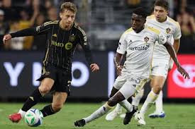 A soccer player in a black and gold jersey dribbles the ball past two players in white jerseys.